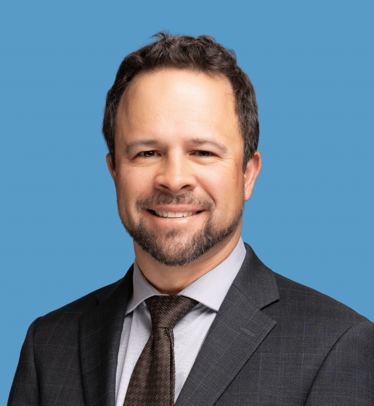 Headshot of Asher David, a smiling man with brown hair and short-haired beard in a grey suite, light great shirt, and tie, against a blue background