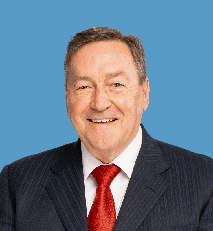 Headshot of Cal Trudeau, a smiling middle-aged man with light brown hair, wearing a dark suit, with a white shirt and red tie, posed against a blue background