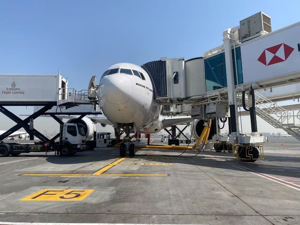 A Boeing 777 aircraft at the airport apron, with airport ground service equipment and signage visible.