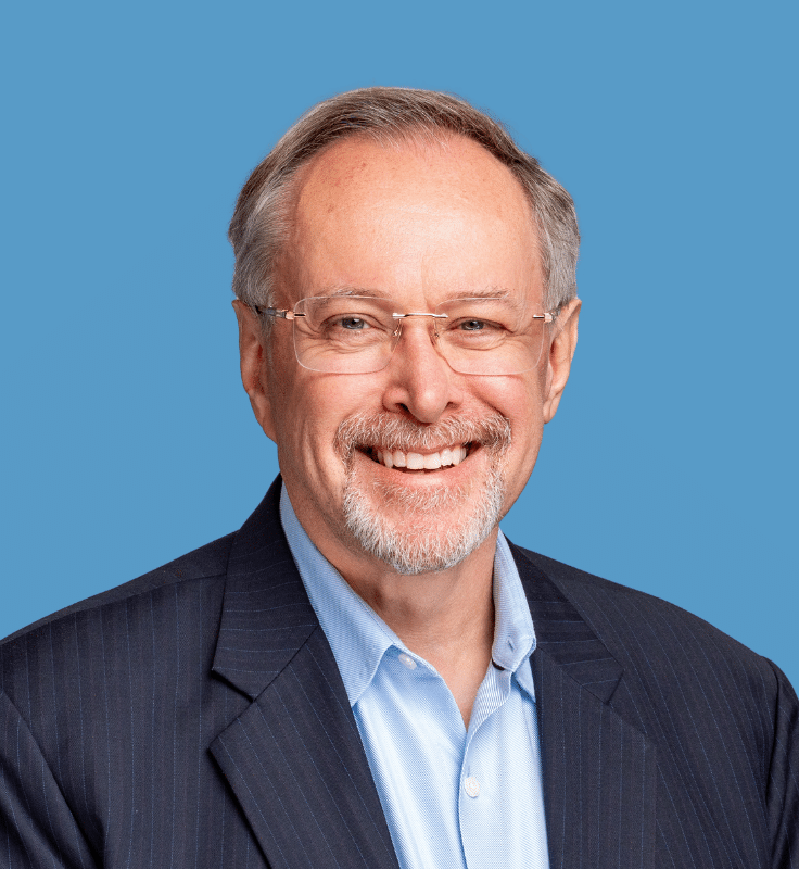 Headshot of Eric Miller, a smiling man with grey hair and a full goatee, wearing rimless glasses, a navy suit with a light blue shirt, against a blue background