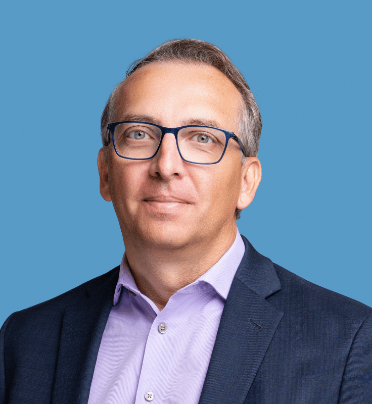 Headshot of Felix Rosbergen, a smiling man wearing glasses, in a navy suit and lavender button-down shirt, against a blue background