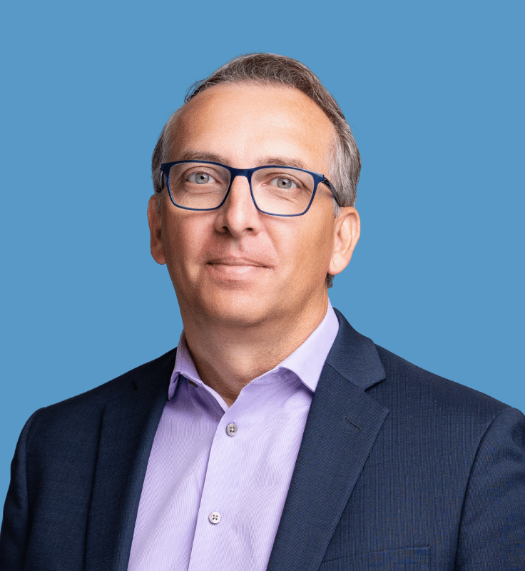 Photo of a man wearing glasses, in a navy suit and lavender button-down shirt