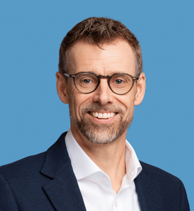 Headshot of Henrik Krogh Hansen, a smiling man with glasses, and light brown hair and beard, wearing a dark navy suit jacket and white button-down shirt, against a blue background