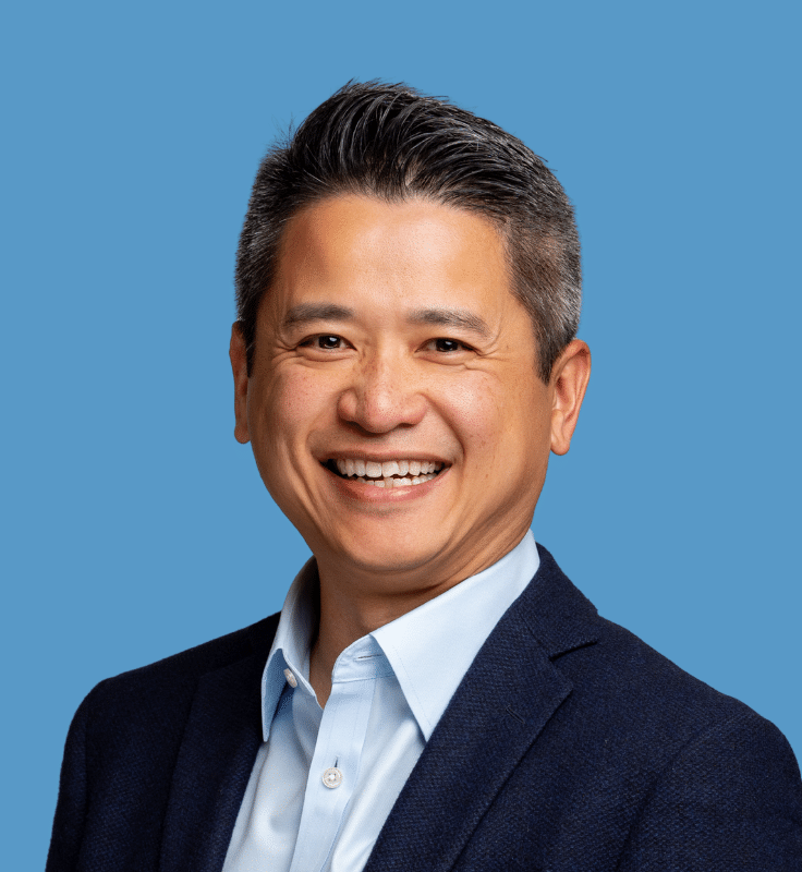 Headshot of John Sum, PMP, a smiling Asian man with black hair, wearing a dark navy suit and light blue button-down shirt, against a blue background