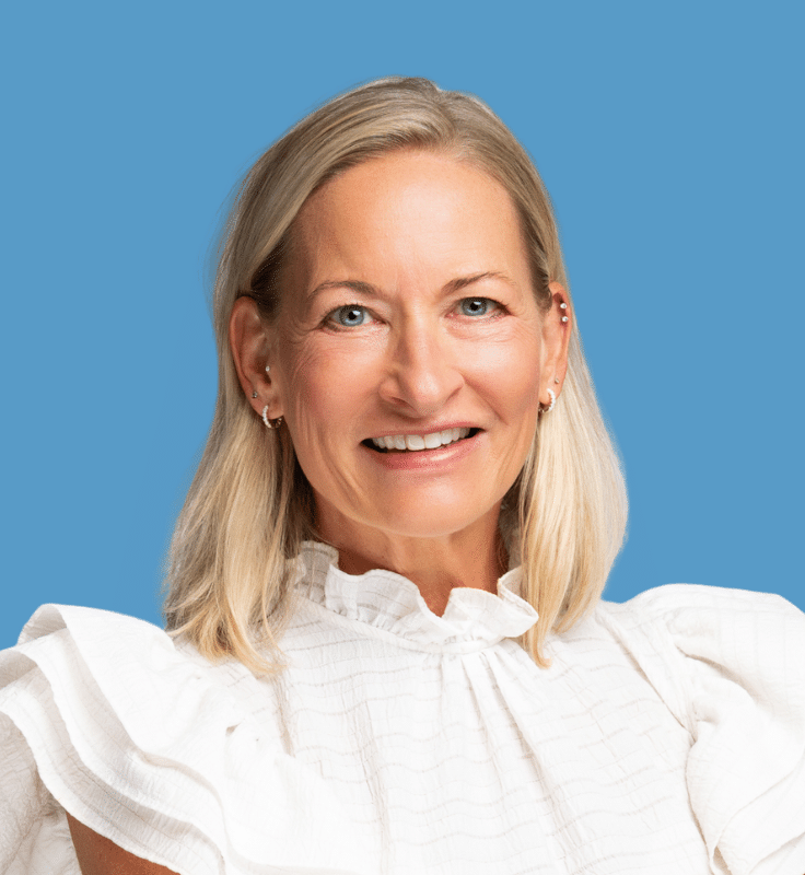 Headshot of Michelle Brown-Daly, a smiling woman with blonde hair wearing a white blouse with ruffles, posed against a blue background