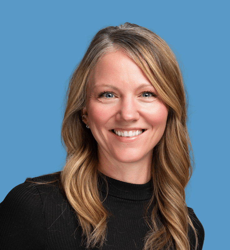 Headshot of Kaley Konecny, a smiling woman with blonde hair wearing a black sweater against a blue background.