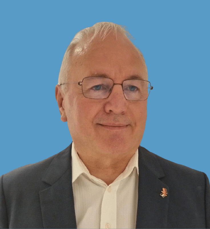 Headshot of Kim Madsen. A man with white hair and glasses, wearing a black suit and white button-down shirt, against a blue background